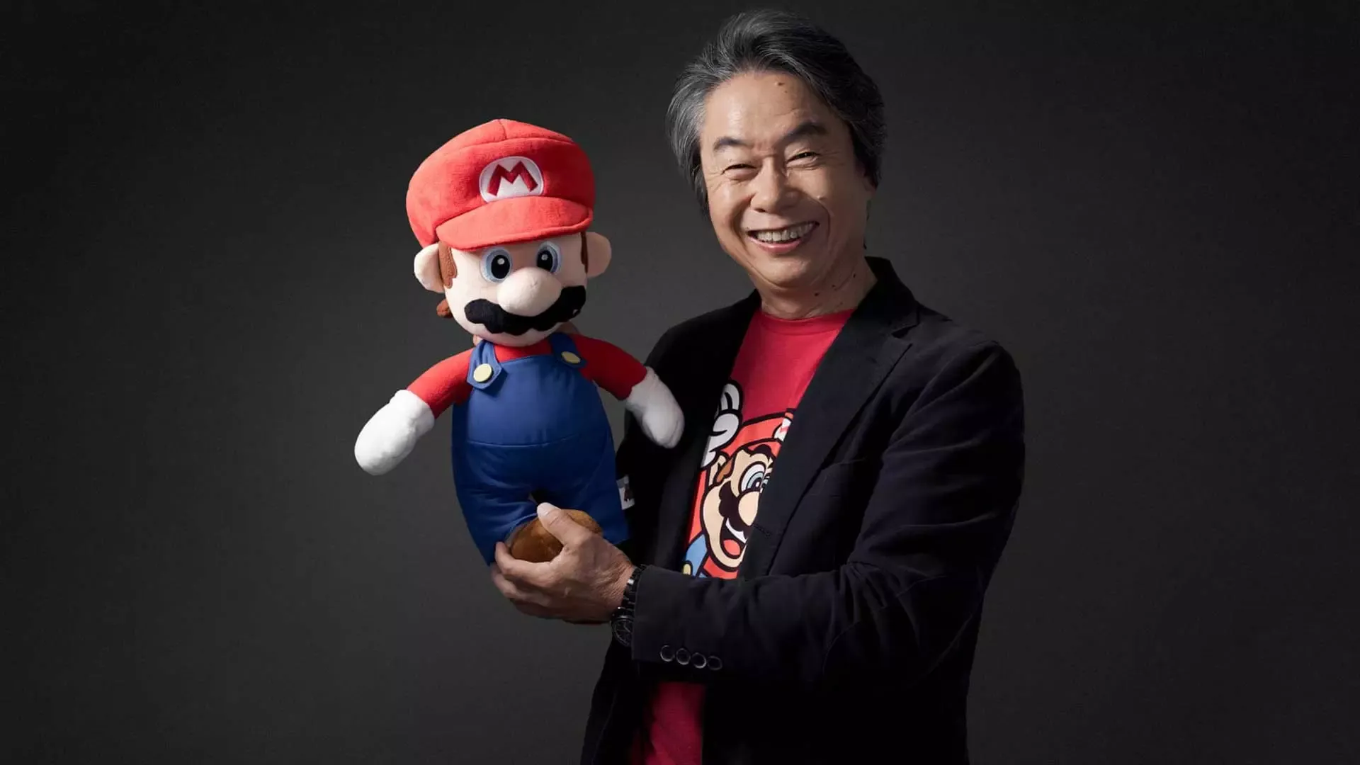 Shigeru Miyamoto sorri enquanto segura um boneco de pelúcia do Mario, diante de um fundo escuro.